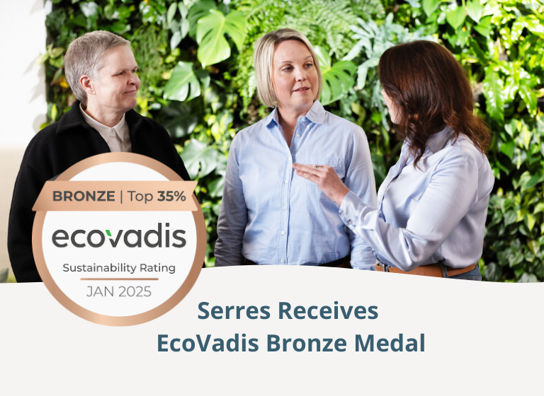 Three women standing in front of a green plant wall having a discussion. EcoVadis bronze medal in front.